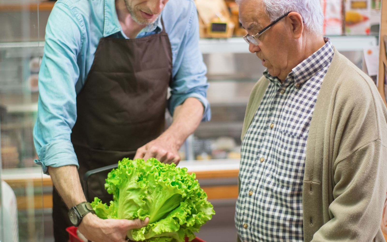 A friendly helper holds fresh lettuce while assisting an older man at a neighborhood grocery.