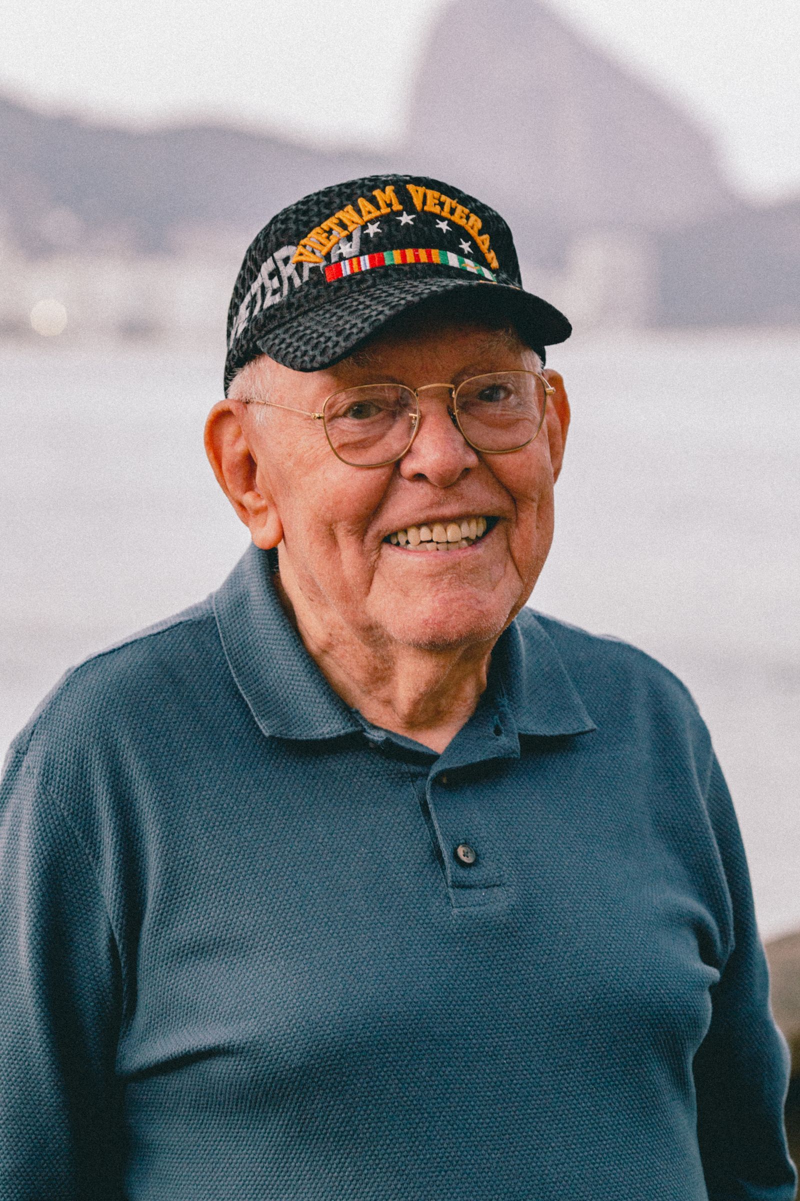 An older Vietnam veteran in a service cap smiles beside the ocean.